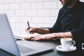 Cropped image of stylish businessman dressed in black shirt making notes in notepad working at...