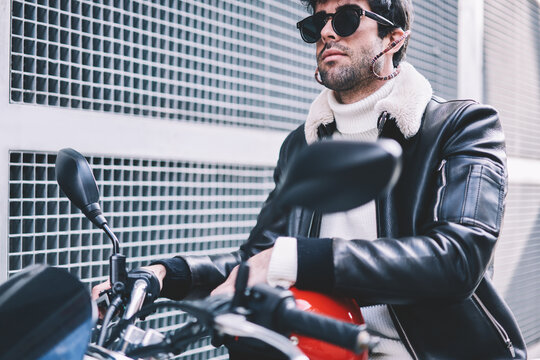 Cropped image of serious bearded male in black sunglasses with helmet sitting on motorcycle in urban setting.Stylish biker dressed in denim jacket driving on motorbike on city street