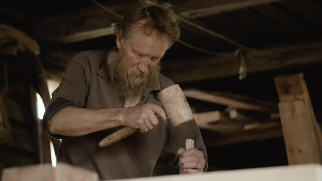Male carpenter with a gray beard, works with a hammer on a tree and chisel. An elderly artisan will carry out his work intently in a workshop. Handwork of the master.