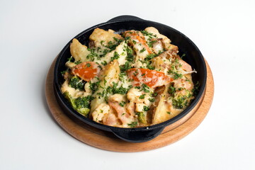 Dish on a black pan, which stands on a wooden stand. Potatoes with cheese and red pepper. Sprinkled with green parsley.