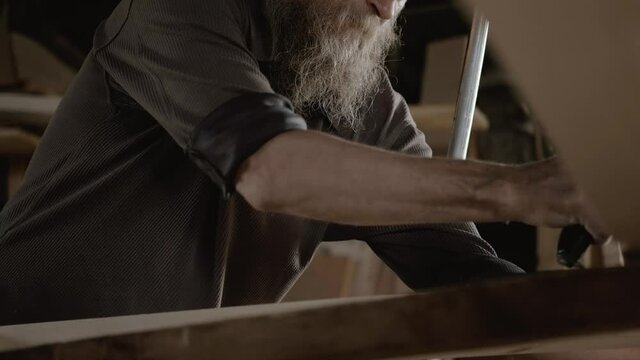 Male carpenter with a gray beard, processes a wood product with a planer. An elderly artisan will carry out his work intently in a workshop. Handwork of the master.