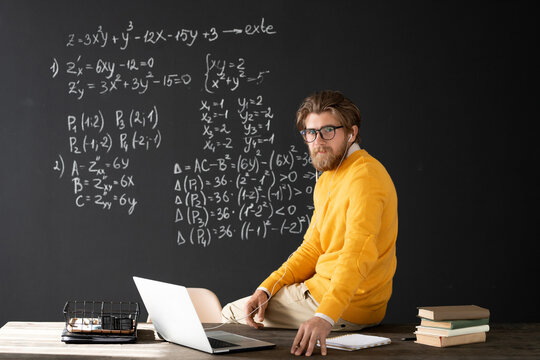 Young serious teacher with earphones looking at you while sitting on table