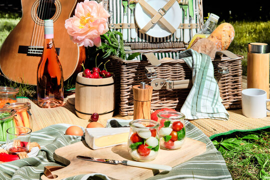 Zero waste picnic al fresco. Vintage picnic basket, hamper with baguette and lemonade outdoors on a grass with cheese, mozzarella, tomatoes, cherries, vine. Guitar on straw blanket and stripy towels.