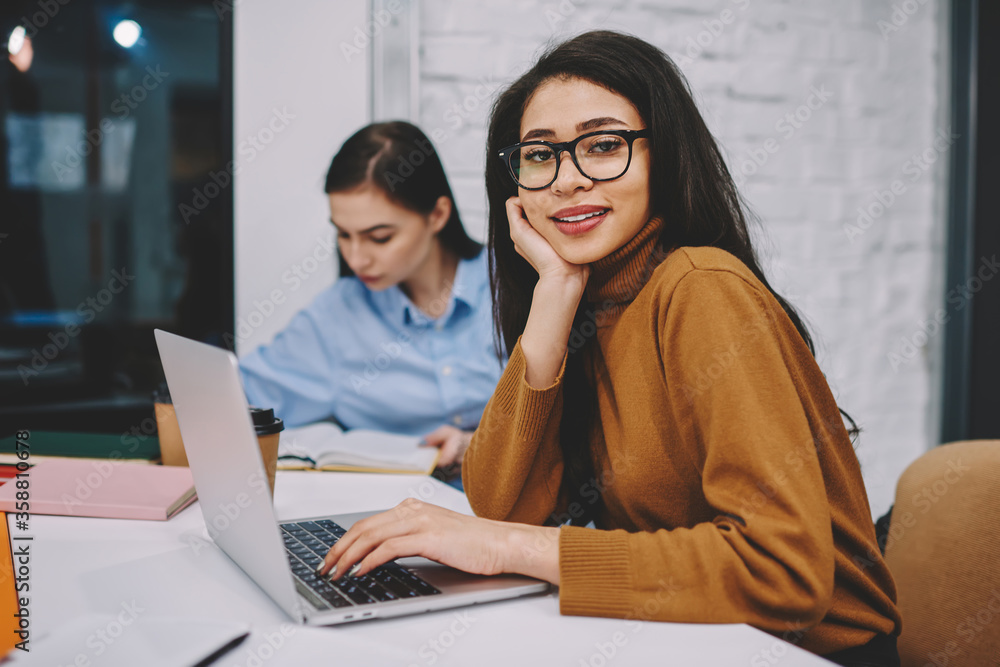 Poster portrait of smiling young woman in spectacles using laptop computer for learning in campus, positive