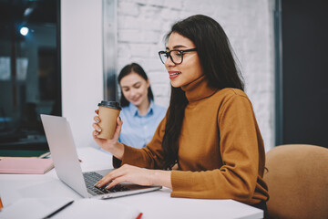 Positive brunette girl in spectacles for vision protection working on laptop computer holding cup of coffee for productivity, smiling young woman watching video online on netbook on break in office.