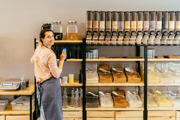 Owner woman with rag cleaning glass dispenser with metal tap with oil. Woman assistant in zero waste shop. Weighing dry goods in plastic free grocery store. Sustainable shopping at local business.