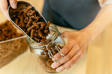 Dark brown whole grains of pasta are poured in glass jar. Close up of unrecognizable woman is pouring pasta to a jar from bulk dry food dispensers at zero waste shop, defocused.