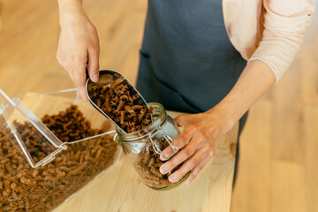 Dark brown whole grains of pasta are poured in glass jar. Close up of unrecognizable woman pouring pasta from plastic free packaging paper bag into storage jar.