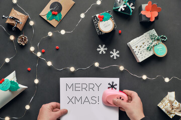 Text Merry Christmas on perforated paper. Presents wrapped in gift boxes with tags. Hands holding paper and puncher. Flat lay, top view with light garland, green alarm, snowflakes on black paper.