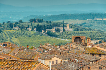 Rooftops of San Gimignano, Tuscan countryside with rolling hills and Santa Maria Assunta a Monte Oliveto Minore church in the distance seen from Parco della Rocca, San Gimignano. - obrazy, fototapety, plakaty