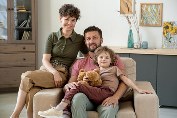 Happy young man with cute little son and his pretty wife sitting in armchair