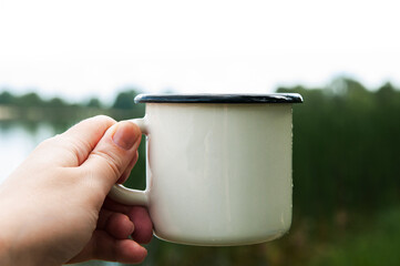 white enameled mug in a hand on a background of nature, calm vacation, place for text