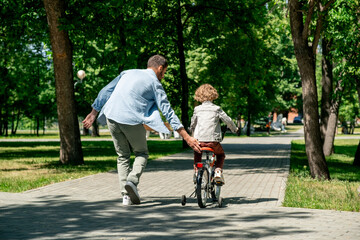 Rear view of young father helping his cute little son ride bicycle outside