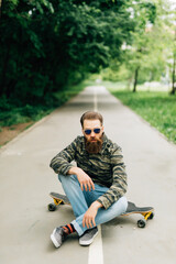 Young bearded man longboarder in casual clothes sitting on the longboard or skateboard outdoors, side view. Urban, subculture, skateboarding concept