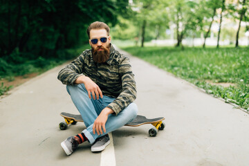 Young bearded man longboarder in casual clothes sitting on the longboard or skateboard outdoors,...