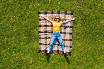 Top view young pretty woman with raised hands lying on the plaid blanket on green grass. - Powered by Adobe