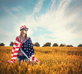 Beautiful young girl holding an American flag in a field of wheat. Summer landscape against the blue sky. Independence Day Fourth of July
