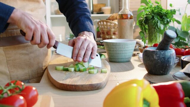 Arc midsection shot of professional male chef in apron chopping fresh cucumber with butcher knife on wooden board while cooking at kitchen table