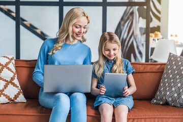 Beautiful young mother and her daughter using gadgets and smiling while sitting on sofa at home