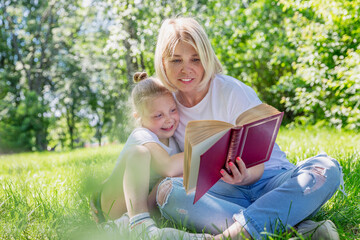 Young blond woman with little daughter lie on the grass with a book in the park. Love and tenderness. Camping on a sunny summer day.