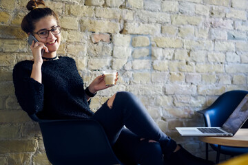 Laughing young woman in optical eyeglasses talking on smartphone with friends while holding tasty aroma coffee in hand and sitting at opened laptop device during recreation time in cafe indoors