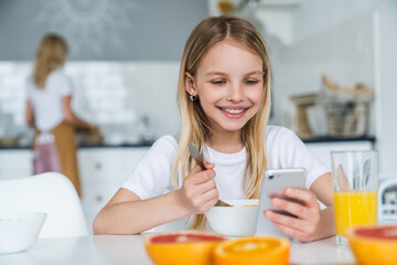 Little girl having breakfast while using phone at home kitchen