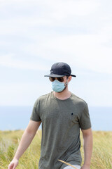portrait of a man with a face mask a cap and glasses in nature with the sea and sky in the background