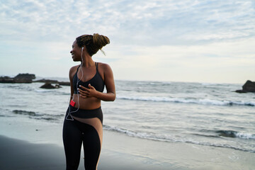 Fototapeta premium Afro american young woman with dreads listening audio songs from own player downloaded on smartphone resting outdoor on coastline, dark skinned jogger enjoying music in earphones connected to cellular
