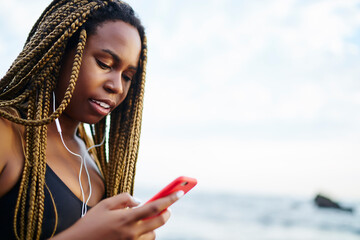 Dark skinned female lover of sport with dreads choosing music songs in mobile app for morning run outdoors, attractive black woman jogger in earphones listening audio from smartphone before workout