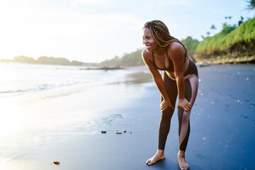 Cheerful athletic afro american jogger dressed in tracksuit has break during morning run along seashore.Positive female with dark skinned happy after hard sport workout on coastline of tropical island