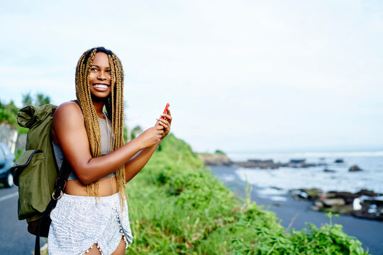 Happy afro american young woman with backpack laughing while making pictures of tropical environment on smartphone.Positive female tourist with dark skin taking photos on cellular travelling on island