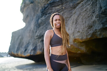 Portrait of cheerful dark skinned sportwoman dressed in active clothes smiling at camera.Positive afro american young woman in tracksuit standing outdoors on coastline before morning workout