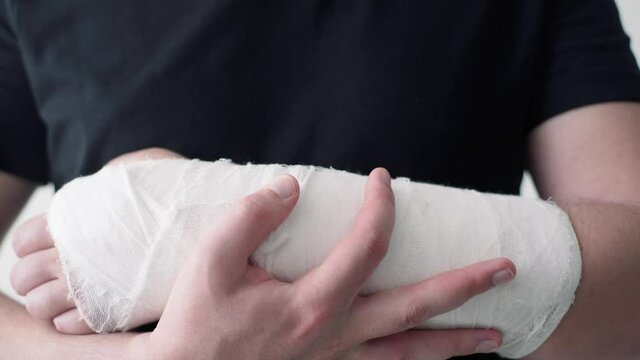 A healthy hand of a young man supports a wounded broken arm in a plaster bandage close-up. The difficulty of self-movement of the hand at a fracture
