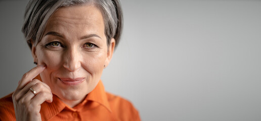 Charming gray-haired woman smiles touching cheek with finger. Selective focus on female face with...