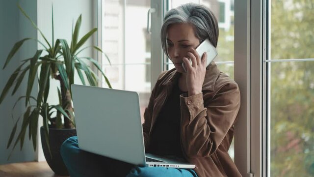 Gray haired mature woman answering phone while working on laptop sitting leaning back against the window. mature people in business. Business concept. Prores 422.