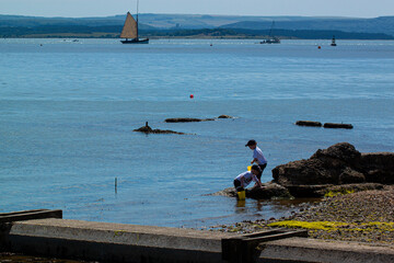 twin boys are playing on a rugged dirty beach with the river and sail boats in the background.