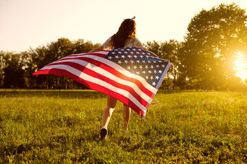 Back view of young woman with American flag running towards sunset in countryside. Independence Day celebration