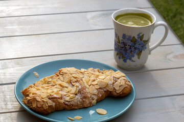 Almond croissant and matcha latte in white cup with flowers standing on light-blue wooden table in sunny summer day. Tasty morning of young trendy person