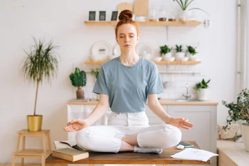  Peaceful redhead young woman is sitting with closed eyes on table in lotus position and makes deep breath-exhalation at home office. Peaceful female sitting in lotus pose. © dikushin