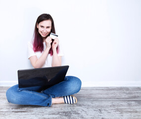 Young woman sitting on floor holding laptop happily smiles and in hands credit card, grey background. Copy space