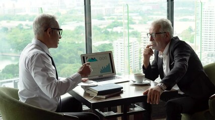 two senior businessman take a break drinking coffee and discussing business strategy on table with graph in laptop computer in coffee shop at modern restaurant on a tall building cityscape background - Powered by Adobe