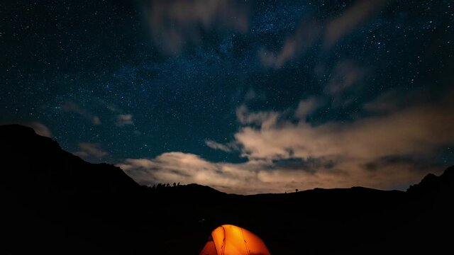Orange tent with a burning light inside stands in a clearing under the starry sky. On the horizon mountains and forest. Concept of a beautiful starry night sky and the Milky Way. Night in the mountain