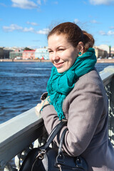 Fototapeta premium Smiling adult woman wearing coat standing on bridge, looking at the distance, sunny weather is in a city