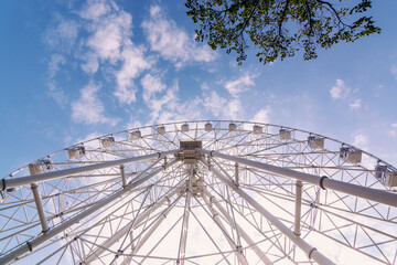 White metal construction of an attraction Ferris wheel with observation cabs high against a blue sky with white clouds.