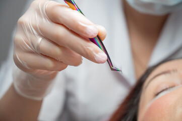 Woman lies on eyelash extension procedure in a beauty salon. Lashmaker holds tweezers with a bunch of artificial eyelashes. Close-up of a craftsman in gloves.