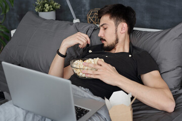 Young man having got popcorn all over him lying with laptop on bed and eating popcorn from tshirt