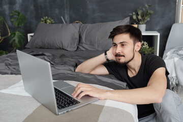 Content young man with mustache using laptop on bed while chatting via video conferencing app during self-isolation