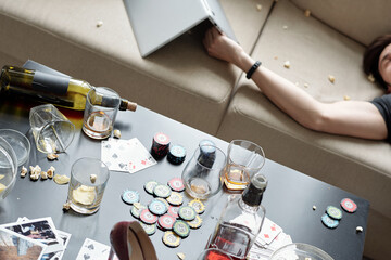 Above view of drunken man sleeping on sofa with laptop after party, messy food and glasses on table with poker chips