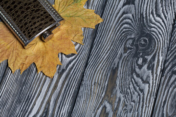 Flask for an alcoholic drink. Autumn maple leaves are yellow. On pine boards painted black and white.