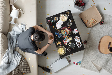 Above view of brunette man sitting on sofa in dirty room with messy bottles and snacks and remembering yesterday party